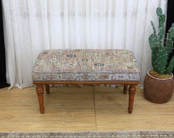 Puede incluir: Un otomán rectangular con una parte superior de tela estampada en tonos rosa, azul y beige. El marco de madera tiene patas torneadas. Un cactus en maceta está a la derecha. El fondo incluye cortinas blancas y una cortina negra.