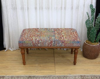 Puede incluir: Un otomán rectangular con una parte superior de tela estampada en tonos rojos, azules y amarillos. El otomán tiene un marco de madera y cuatro patas torneadas. La pieza es un mueble decorativo.