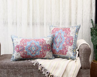 May include: Two decorative throw pillows with a vintage rug design in red, blue, and cream colors. One pillow is rectangular, and the other is square. They rest on a brown patterned bench with a cream-colored fringed throw blanket.