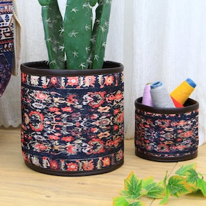 Könnte beinhalten: Zwei zylindrische Aufbewahrungskörbe mit einem farbenfrohen Muster. Die Körbe sind aus Stoff und haben einen braunen Rand. Der größere Korb ist mit einer Kakteenpflanze gefüllt. Der kleinere Korb ist mit bunten Garnen gefüllt.