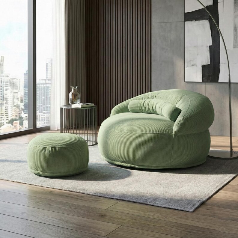 Pode incluir: Uma cena de sala de estar moderna com uma poltrona de veludo cotel&ecirc; verde claro e um pufe a condizer. A cadeira arredondada tem encosto e apoio de bra&ccedil;os curvos. O pufe &eacute; circular. A sala tem uma janela grande, uma mesa lateral e um tapete neutro.