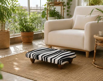 May include: A rectangular footstool featuring a bold black and white zebra stripe pattern. The low-profile piece is supported by four small, rounded wooden feet, providing a stable base for resting feet while seated in a living room environment.