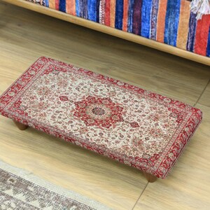 Peut inclure: Un repose-pieds rectangulaire avec un motif floral détaillé en rouge, crème et marron. Le repose-pieds a une bordure rouge et est soutenu par quatre petits pieds en bois. Le design rappelle un tapis traditionnel.