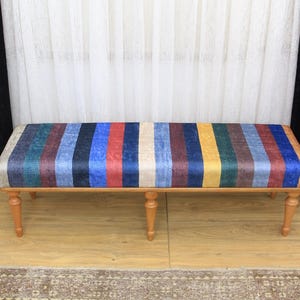 May include: A wooden bench with a colorful striped fabric seat. The fabric is a mix of blue, red, yellow, and green stripes. The bench has four legs and is sitting on a wooden floor.