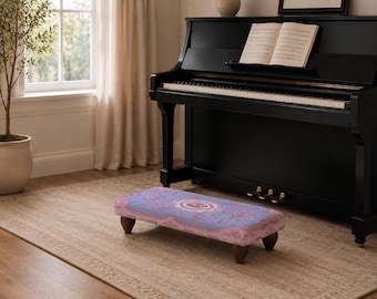 Op de afbeelding: Een rechthoekige pianovoetenbank met een lichtpaarse stoffen bovenkant met een gedetailleerd roze en wit bloemenpatroon. De voetenbank wordt ondersteund door vier korte, donkerbruine houten poten en staat op een lichtgekleurd vloerkleed.