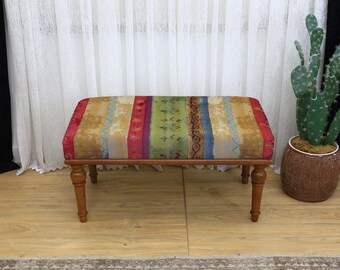 May include: A rectangular upholstered bench with a colorful striped pattern in red, gold, green, and blue. The bench has a wooden frame and four turned legs. A decorative cactus plant in a brown pot is to the right.