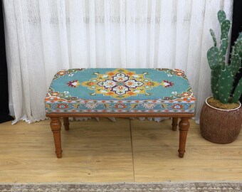 Può includere: Un pouf rettangolare con un piano fantasia turchese e gambe in legno. Il motivo presenta un medaglione centrale con accenti arancioni, gialli e viola. Le gambe sono di colore marrone scuro e il pouf è posto su un pavimento in legno.