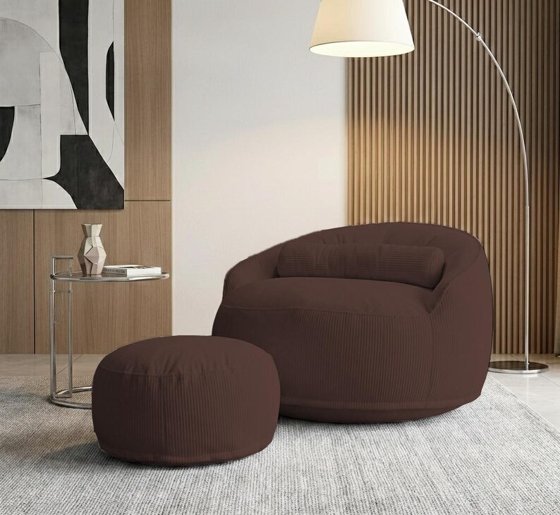 Pu&ograve; includere: Una poltrona a sacco in velluto a coste marrone con un pouf abbinato. La sedia ha lo schienale arrotondato e un cuscino rettangolare. Il pouf &egrave; circolare. I mobili sono su un tappeto grigio.