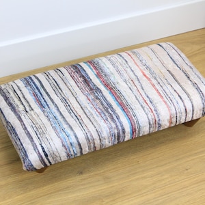 May include: A striped footstool with a beige, blue, red, and black pattern. The footstool has wooden legs.
