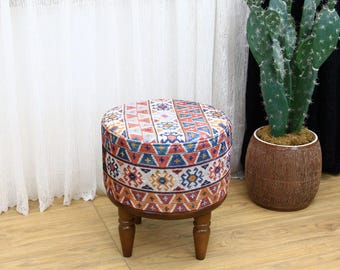May include: A round, patterned ottoman with a wooden base and four legs. The ottoman is covered in a colorful fabric with geometric designs in shades of red, blue, orange, and white. The ottoman is sitting on a wooden floor.