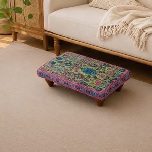 Könnte beinhalten: Ein rechteckiger, niedriger Fußhocker mit einer leuchtend grünen Stoffoberfläche, die ein mehrfarbiges Blumenmuster in Blau-, Rosa- und Violetttönen aufweist. Der Hocker steht auf vier kurzen, dunkelbraunen Holzbeinen und hat eine Zierborte.