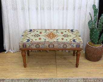 Può includere: Un pouf rettangolare con struttura e gambe in legno. La parte superiore imbottita presenta uno sfondo color crema con un motivo floreale dettagliato nei toni dell'arancione, del blu e del marrone. Le gambe sono di colore marrone caldo. Misura circa 90 cm di lunghezza.