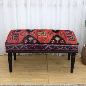 Può includere: Un pouf rettangolare con un top fantasia nei toni del rosso, blu navy e crema, sostenuto da quattro gambe tornite nere. Il design del tessuto presenta motivi geometrici e floreali. Il pouf è un elemento d'arredo decorativo.
