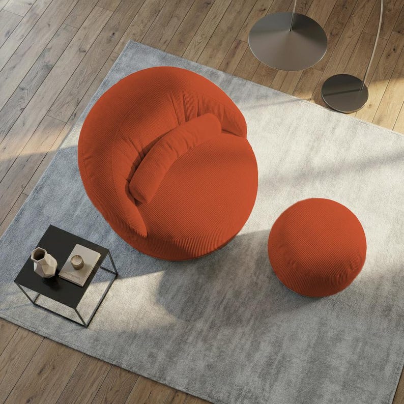 Pode incluir: Uma poltrona redonda laranja com um pufe a condizer est&aacute; sobre um tapete cinzento. Uma pequena mesa lateral preta com objetos decorativos est&aacute; no canto. A cadeira tem encosto curvo e uma pequena almofada presa.