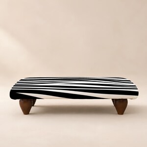 Peut inclure: Un pouf rectangulaire arborant un motif rayé noir et blanc inspiré du zèbre sur sa surface supérieure. Le meuble est soutenu par quatre pieds courts et arrondis en bois brun foncé qui assurent une base stable à ce siège bas.