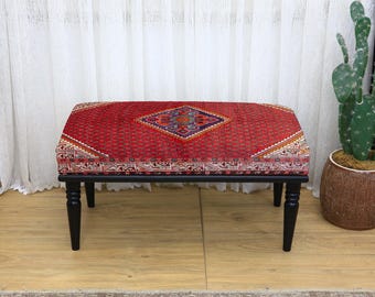 Puede incluir: Un otomán rectangular con una parte superior roja estampada y patas negras. La tela roja presenta un diseño de diamante central con detalles en azul, morado y naranja. El otomán es un mueble decorativo.