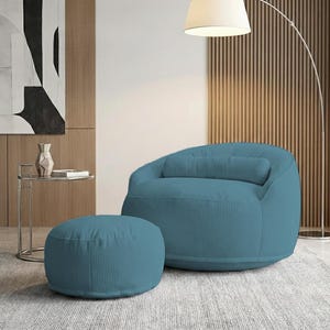 Pu&ograve; includere: Una poltrona in velluto a coste color teal con un pouf abbinato. La poltrona ha una forma arrotondata con un cuscino lombare. Il pouf &egrave; circolare. Una moderna lampada da terra e arte astratta sono sullo sfondo.