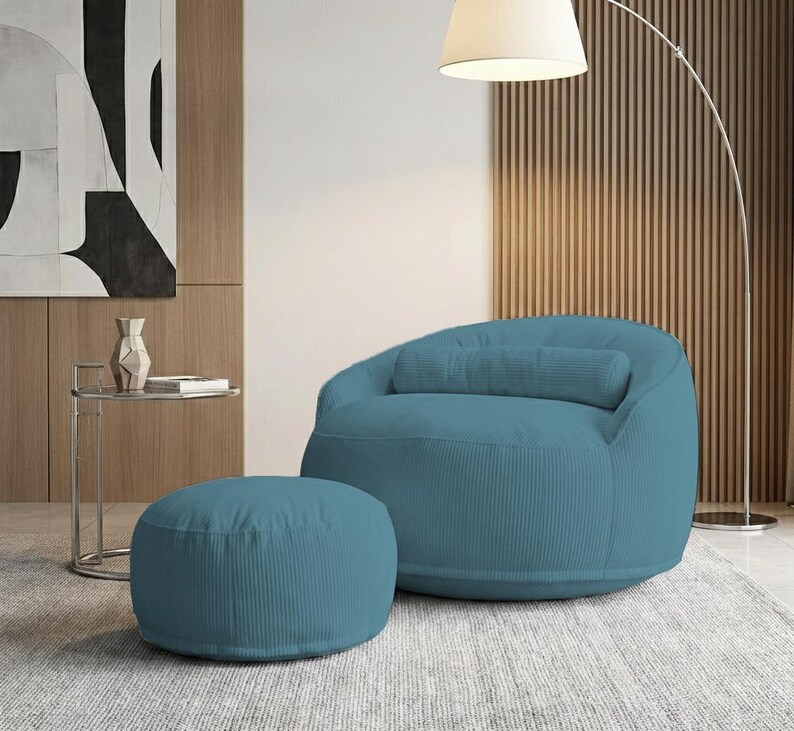 Pu&ograve; includere: Una poltrona in velluto a coste color teal con un pouf abbinato. La poltrona ha una forma arrotondata con un cuscino lombare. Il pouf &egrave; circolare. Una moderna lampada da terra e arte astratta sono sullo sfondo.