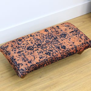May include: A small, rectangular footstool with a patterned fabric top. The fabric is a mix of orange and black with a floral design.