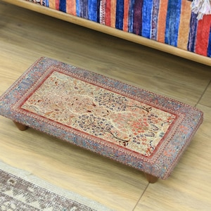 Peut inclure: Un pouf rectangulaire bas avec un dessus à motifs détaillés. Le motif présente un motif floral central dans des tons de rouge, beige et bleu, encadré par une bordure rouge et un bord à motifs. Le pouf repose sur quatre petits pieds en bois.