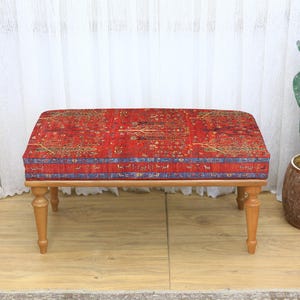 May include: A rectangular ottoman with a red patterned top and light brown wooden legs. The red fabric has a detailed design with blue accents. The ottoman is a low, bench-style seat.