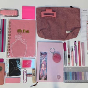Op de afbeelding: Een assortiment roze en witte briefpapier en accessoires, waaronder een notitieboek, pennen, stickers, een etui en een paar fluffy sokken met hartpatronen. Een pot met een bloemenontwerp is ook zichtbaar.
