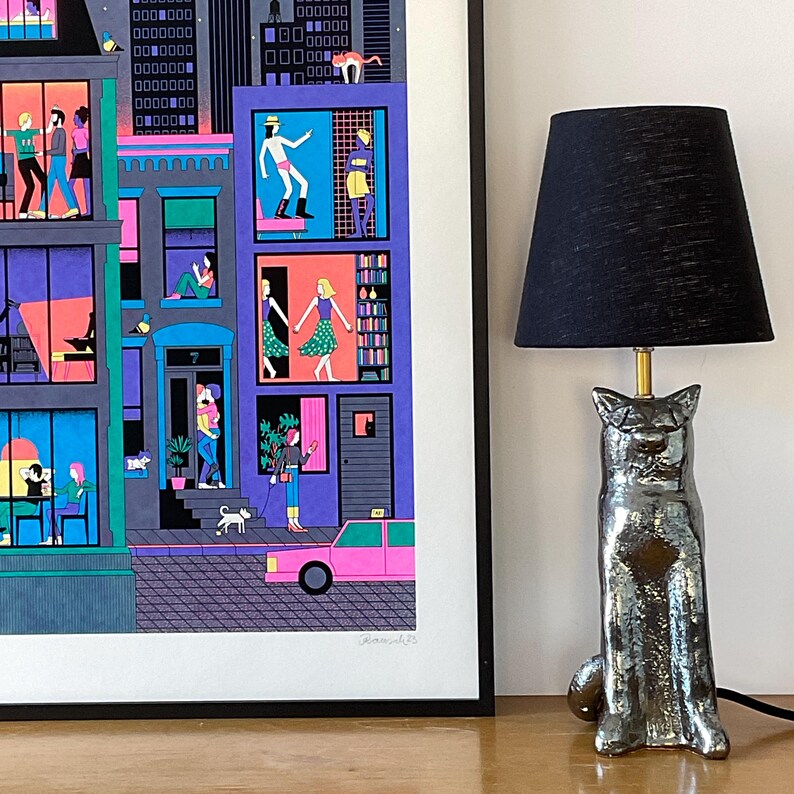 May include: A framed print of a colorful city scene with people in windows and a car driving down the street. A black lamp with a cat-shaped base sits on a wooden table in front of the print.