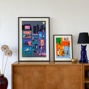 May include: Two framed illustrations of city scenes. The first illustration depicts a multi-story building with windows showing people inside. The second illustration shows a cafe with people sitting at tables outside. Both illustrations are in a colorful, graphic style.