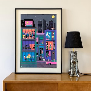 May include: A colorful illustration of a city street at night with multiple apartment buildings. Each window shows a different scene of people interacting inside their homes. The illustration is framed in black and displayed on a wooden shelf with a black lamp.