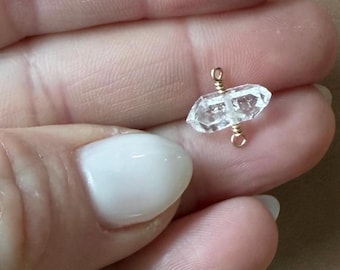 Op de afbeelding: Close-up van kleine, heldere kwartskristallen kralen met goudkleurige draadwikkeling. De kristallen worden in een hand gehouden, met andere vergelijkbare kralen zichtbaar op de achtergrond. De kralen zijn waarschijnlijk voor het maken van sieraden.