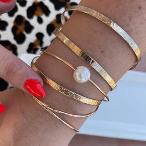 May include: A collection of gold-toned bracelets adorns a wrist. The bracelets include hammered metal bangles and delicate wire designs. A single bracelet features a white pearl. The background includes a leopard print pattern.