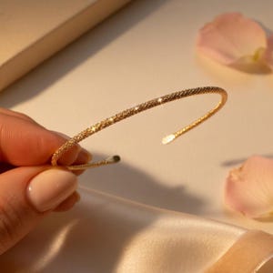 May include: A gold-colored open cuff bracelet with a textured surface. The bracelet is held by a hand and is set against a soft background with pink flower petals.