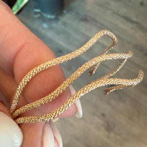 May include: A gold-colored, textured bracelet with a three-strand design. The bracelet has a coiled, open design, and the surface has a granular texture. The jewelry is likely made of metal and is designed to be worn on the wrist.