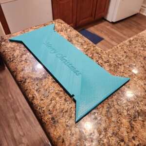 May include: A turquoise-colored, rectangular Christmas decoration with a pointed end on each side. The words "Merry Christmas" are embossed on the surface. The decoration is resting on a speckled countertop.