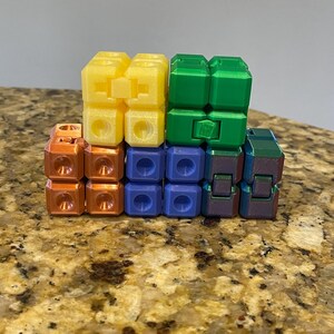 May include: A colorful fidget toy composed of interconnected cubes in yellow, green, blue, orange, and iridescent colors. The cubes are arranged in a stacked configuration, showcasing their geometric design and the variety of hues.