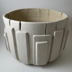 May include: A white ceramic planter with a geometric design. The planter has a wide, open top and a series of rectangular shapes around the exterior. The interior is a lighter shade of white. The planter is suitable for indoor or outdoor use.