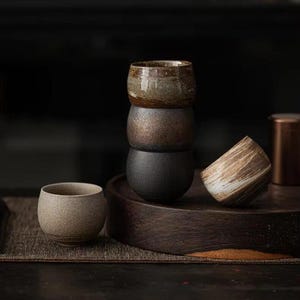 Puede incluir: Un juego de cuatro tazas de té de cerámica apiladas sobre una superficie de madera. Las tazas son de varios tonos de marrón y gris, con un acabado texturizado.