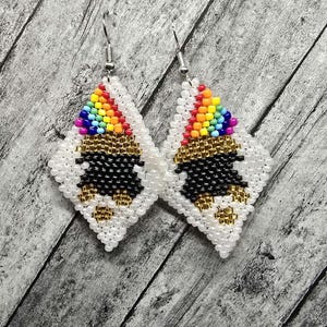 Op de afbeelding: Een paar diamantvormige oorbellen van kralen. De oorbellen hebben een regenboogontwerp aan de bovenkant, met zwarte en gouden accenten. De oorbellen zijn wit met een zilveren haak.