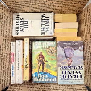 Könnte beinhalten: Ein Karton voller alter Bücher. Titel sind unter anderem "The Seven Minutes" und "Battlefield Earth". Die Bücher sind unterschiedlich groß und farbig, einige weisen Altersspuren auf.