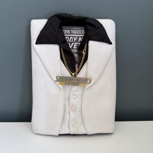 May include: A white jacket with a black collar and a gold chain necklace. The necklace has a pendant that reads "Saturday Night Fever". The jacket is buttoned up and has a movie poster visible in the collar.