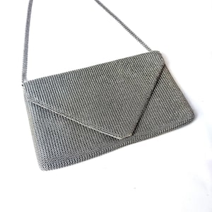 May include: A silver-colored, envelope-style clutch with a chain strap. The bag is made of a woven material, and the flap closure is triangular. The chain strap is also silver.
