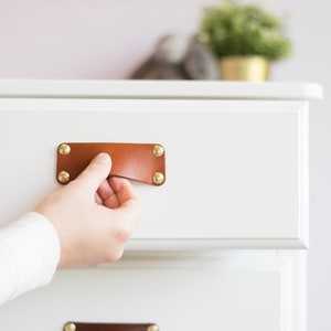 Könnte beinhalten: Eine weiße Kommode mit zwei Schubladen. Die obere Schublade hat einen braunen Ledergriff mit Messingnieten. Die untere Schublade hat einen ähnlichen Griff.