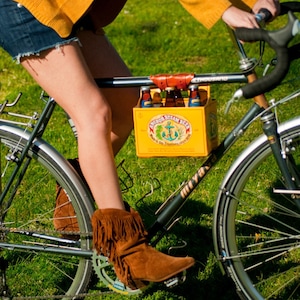 Woman riding a bicycle on grass. A yellow cardboard beer carton is attached to the top tube with the honey leather 6-pack frame cinch.