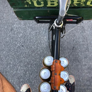 Customer photo looking down on the top tube of a bicycle from the saddle with their feet on the pedals. The street below is in motion and the top tube is holding a 6-pack of beer with a leather carrying strap