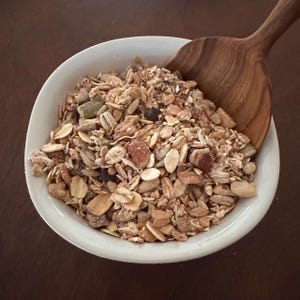 May include: A white ceramic bowl filled with a mixture of rolled oats, various nuts, seeds, and dried fruit pieces. A wooden spoon with a visible grain pattern is partially submerged in the granola mixture, resting against the side of the bowl.