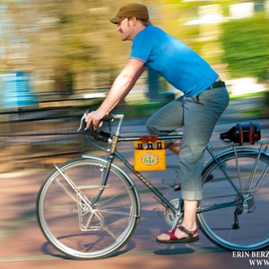 A man riding a bike with the background blurred. A yellow cardboard beer carton is attached to the top tube with the honey leather 6-pack frame cinch.