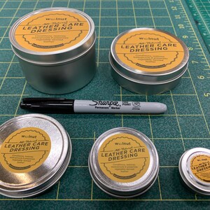 May include: Five silver tins of leather care dressing with yellow labels that say "Walnut All-Natural Leather Care Dressing" and a black Sharpie permanent marker on a green cutting mat.