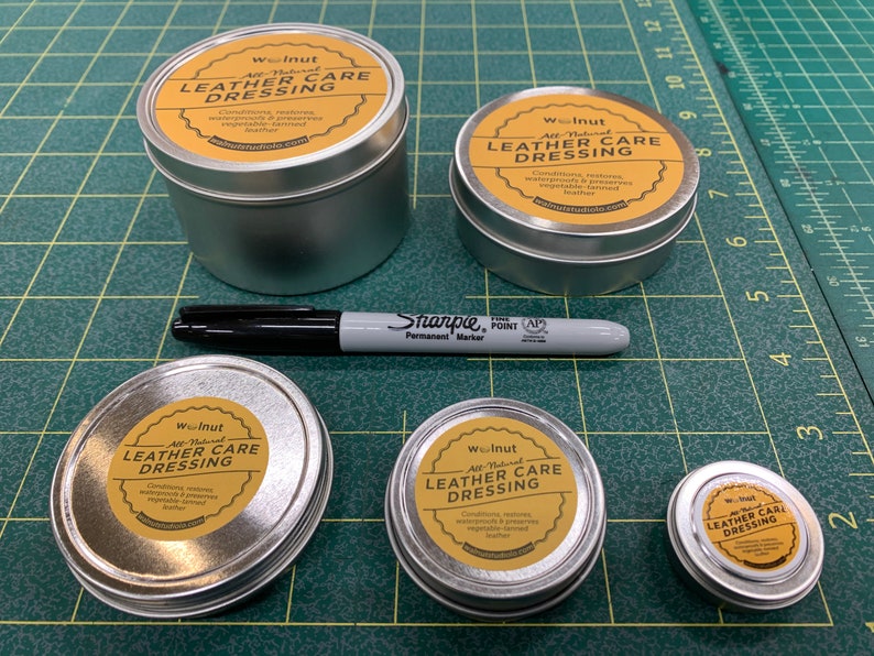 May include: Five silver tins of leather care dressing with yellow labels that say "Walnut All-Natural Leather Care Dressing" and a black Sharpie permanent marker on a green cutting mat.