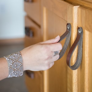 May include: Close-up of two brown leather cabinet pulls attached to a light wood cabinet. The pulls are loop-shaped and secured with silver-colored hardware. One hand is reaching for a pull, with a silver bracelet visible.
