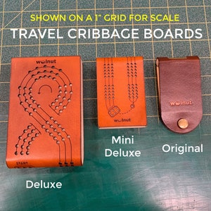 Shows all 3 designs of. Walnut Studiolo's folding leather and wood cribbage boards on a 1-inch grid mat for a size comparison: the Deluxe, Mini Deluxe, and Original Travel Cribbage Boards.